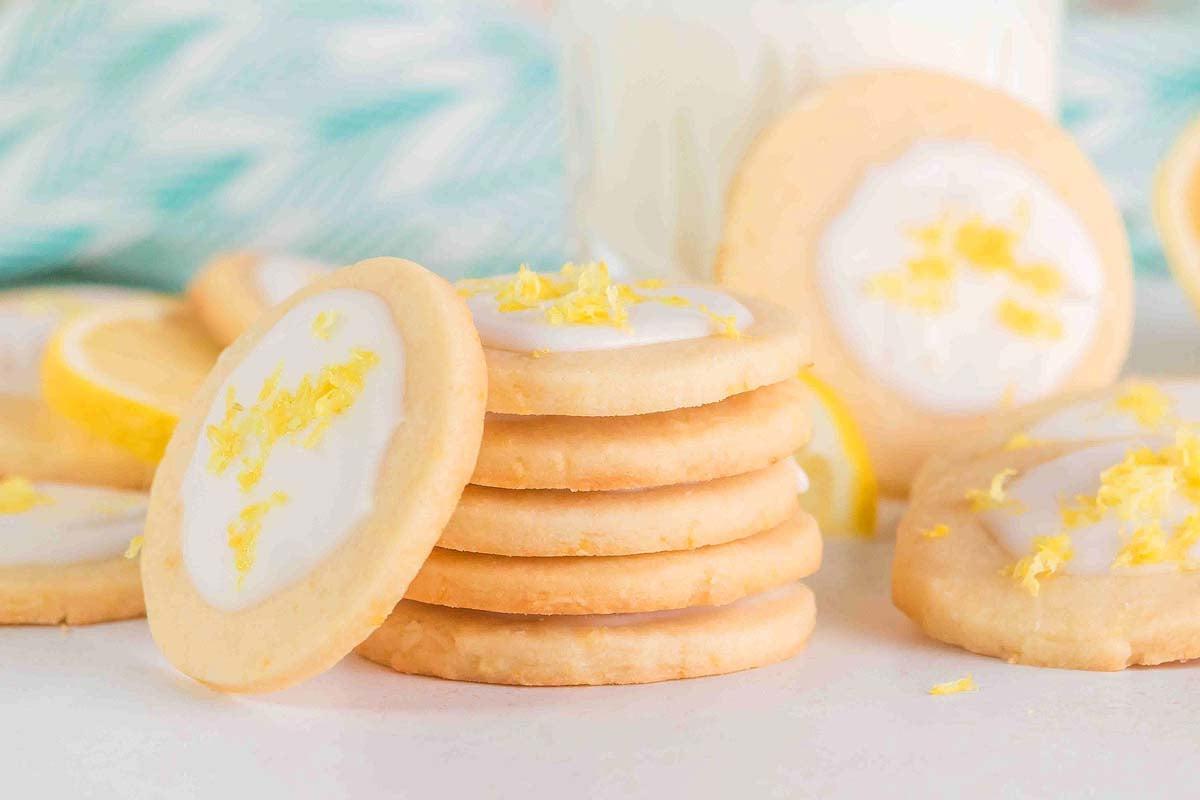 Μια στοίβα από λεμονάτα μπισκότα shortbread με λευκό γλάσο και ξύσμα λεμονιού, περιτριγυρισμένα από περισσότερα μπισκότα και μια υποψία λεμονιών στο παρασκήνιο.