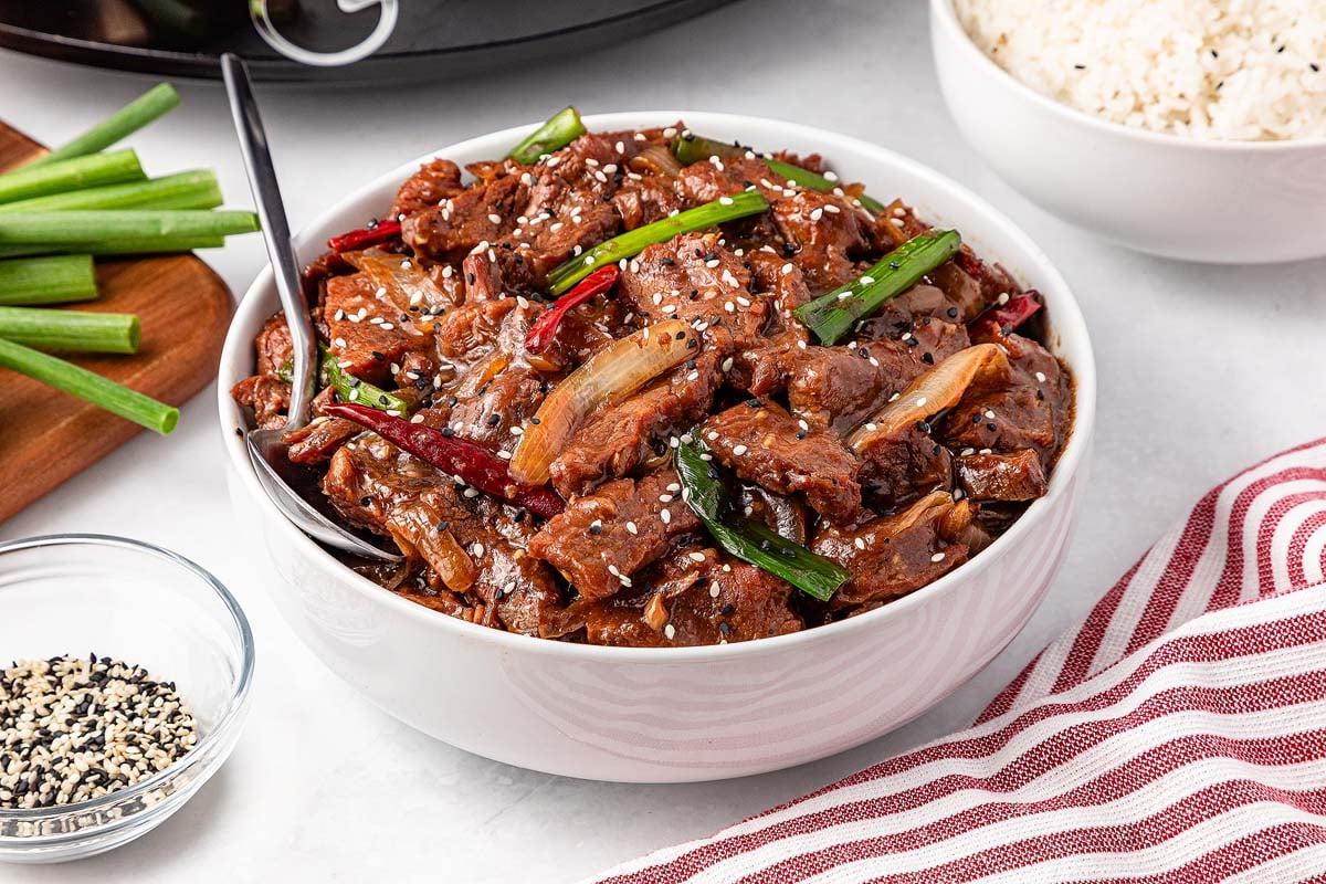 **Σιγομαγειρεμένο Μογγολικό Βοδινό** 2 A bowl of slow cooker Mongolian beef garnished with green onions and sesame seeds sits enticingly on the table, accompanied by a bowl of rice, extra chopped green onions, and a dish of sesame seeds.