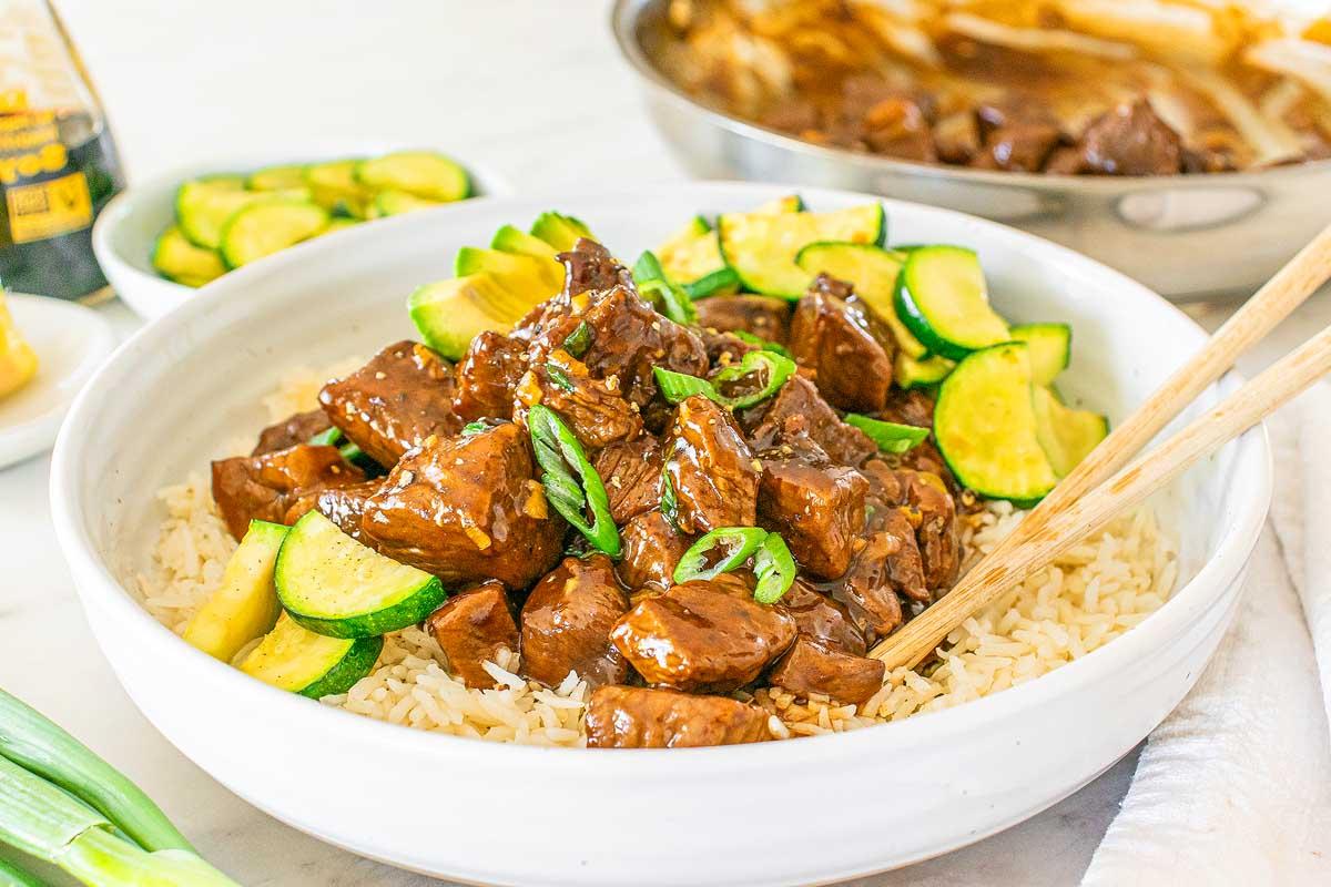 Τεριγιάκι Στέικ Μπουκίτσες 2 Μια μπολ stir-fry τεριγιάκι μοσχαριού με κολοκυθάκια σερβιρισμένο πάνω σε ρύζι, γαρνιρισμένο με φρέσκα κρεμμυδάκια, βρίσκεται φιλόξενα πάνω σε ένα τραπέζι δίπλα σε ένα τηγάνι και ένα μικρό πιάτο με κολοκυθάκια σε φέτες.