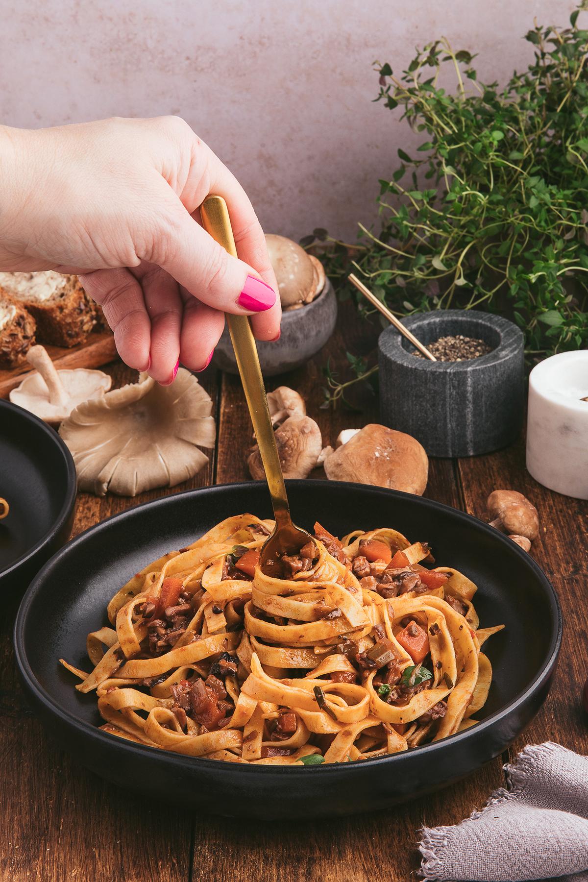 Ραγκού Μανιταριών 11 Ένα χέρι περιστρέφει fettuccine ζυμαρικά με Ραγού Μανιταριών και φρέσκα βότανα σε ένα μαύρο μπολ πάνω σε ένα ρουστίκ ξύλινο τραπέζι.