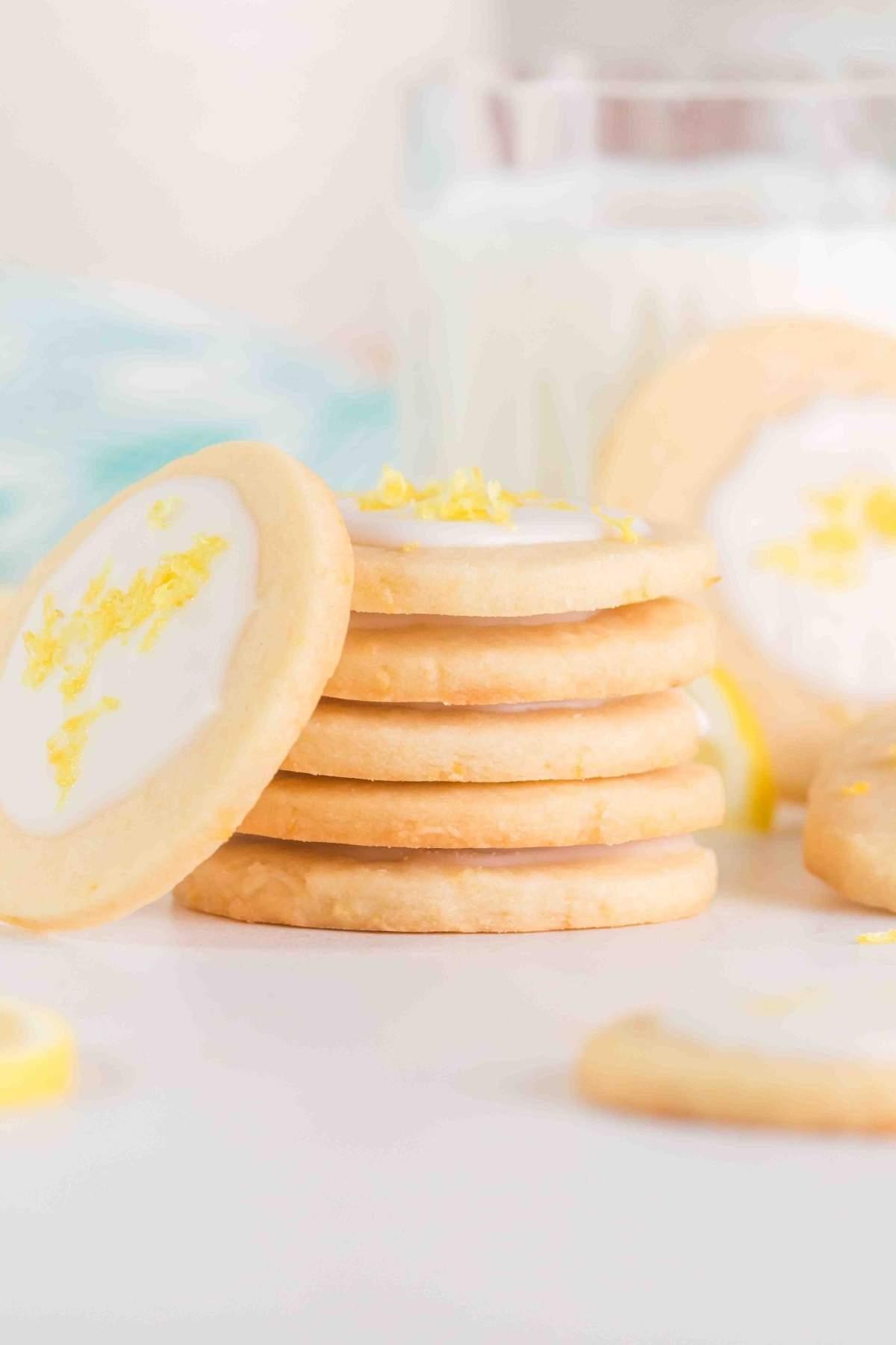 Μια στοίβα από λεμονάτα μπισκότα shortbread με λευκό γλάσο και ξύσμα λεμονιού στην κορυφή, περιτριγυρισμένα από σκορπιστές λιχουδιές. Ένα ποτήρι γάλα βρίσκεται στο παρασκήνιο, έτοιμο να συμπληρώσει την εσπεριδοειδή απόλαυση.