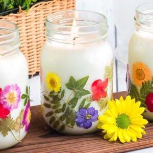 Τρία αναμμένα χειροποίητα κεριά σε βάζο με αποξηραμένα λουλούδια σε γυαλί, το καθένα διακοσμημένο με πολύχρωμα άνθη, τοποθετημένα κομψά σε ένα ξύλινο τραπέζι.