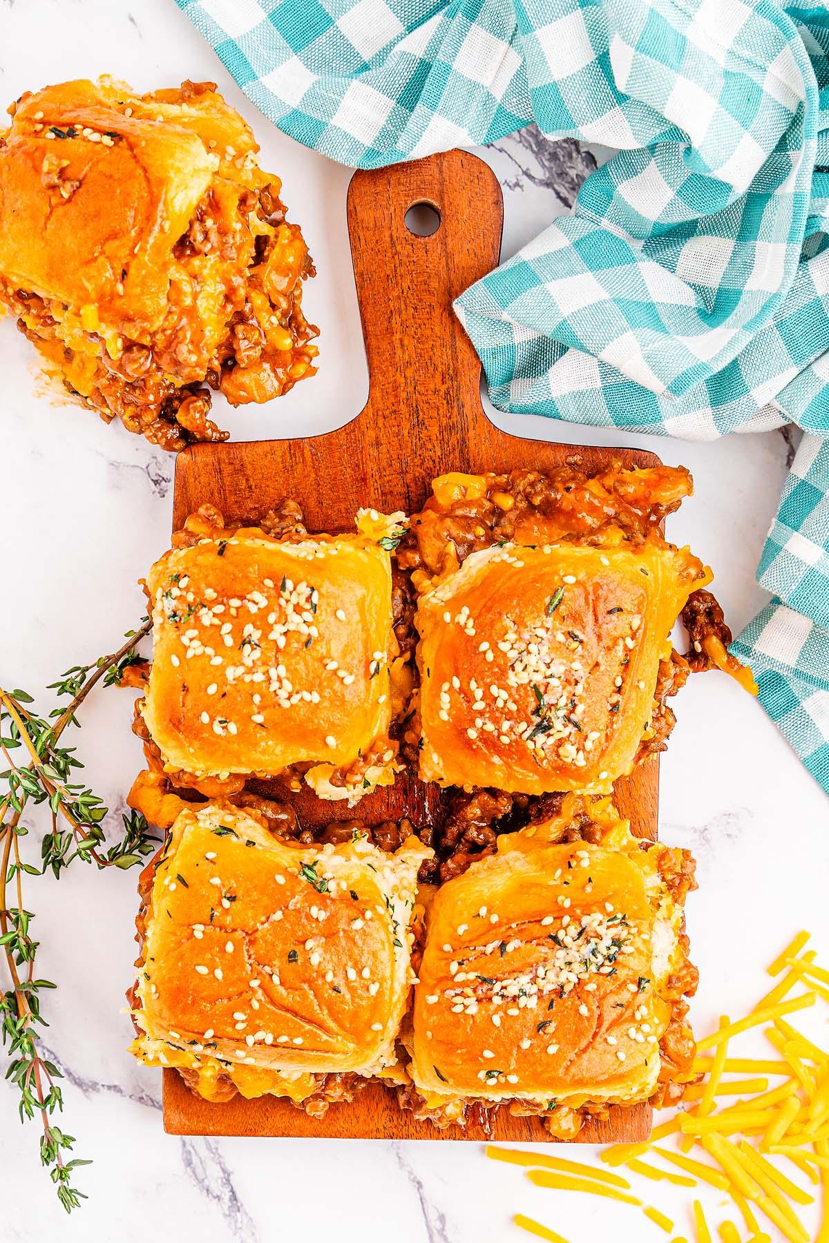 Σλόπι Τζο Μίνι Σάντουιτς 11 Τέσσερα ψημένα Sloppy Joe sliders με μοσχάρι και τυρί σε ψωμάκια με σουσάμι, παρουσιασμένα σε ξύλινη σανίδα και γαρνιρισμένα με μυρωδικά. Τριμμένο τυρί και μια καρό πετσέτα βρίσκονται κοντά.