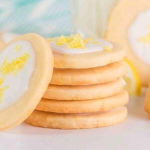 Μια στοίβα από λεμονάτα μπισκότα shortbread με λευκό γλάσο και ξύσμα λεμονιού στην κορυφή ξεκουράζεται κομψά σε μια ανοιχτόχρωμη επιφάνεια, τονισμένη από ένα εν μέρει ορατό μπλε-μοτίφ ύφασμα στο παρασκήνιο.