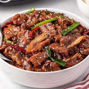 **Σιγομαγειρεμένο Μογγολικό Βοδινό** 12 A bowl of stir-fried slow cooker Mongolian beef with green onions and sesame seeds rests on a white surface, accompanied by rice and chopped green onions.