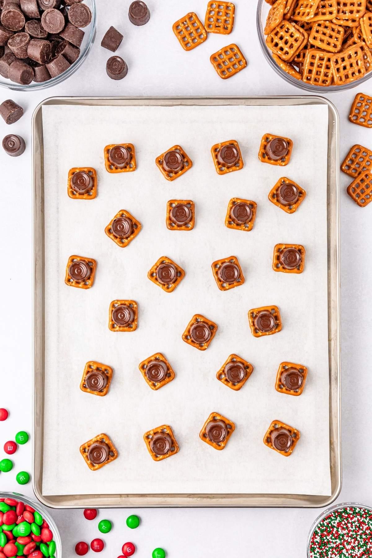 Χριστουγεννιάτικες μπουκιές Rolo pretzel τοποθετημένες σε ταψί, μαζί με μπολ με πρέτσελ, κομμάτια σοκολάτας, ζωντανές κόκκινες και πράσινες καραμέλες M&M και τρούφες.