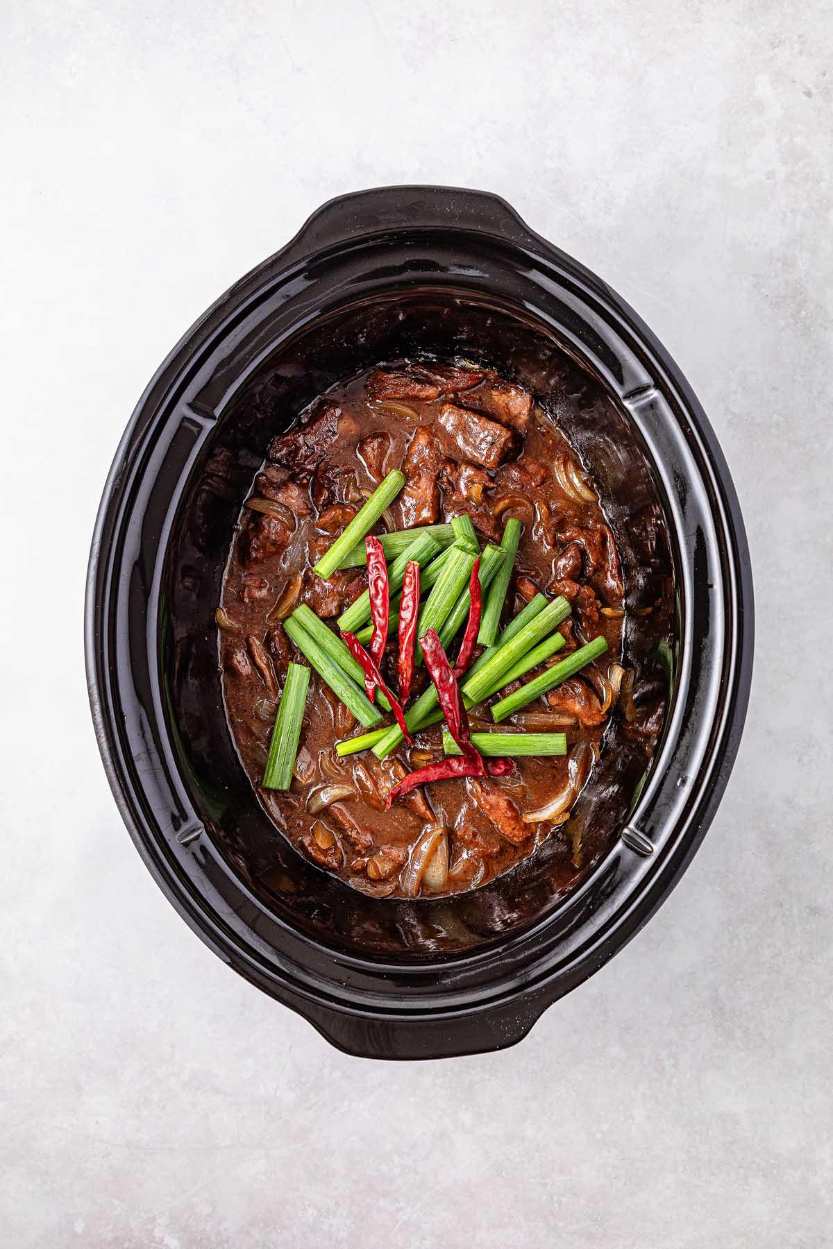 **Σιγομαγειρεμένο Μογγολικό Βοδινό** 17 Slow cooker Mongolian beef stew topped with green onions and red chili peppers, elegantly presented on a pristine white surface.