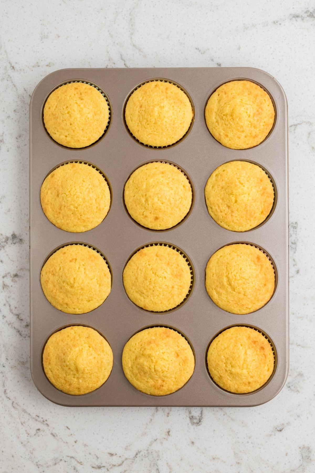 Πασχαλινά Κάπκεϊκς Πουλάκια 17 Ένα ταψί για muffins με δώδεκα φρεσκοψημένα, χρυσαφένια muffins καλαμποκιού σε μαρμάρινο πάγκο.