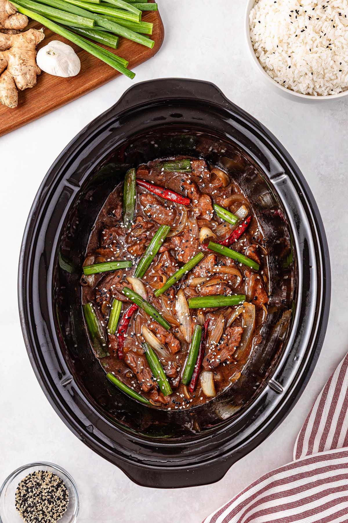 **Σιγομαγειρεμένο Μογγολικό Βοδινό** 18 The slow cooker Mongolian beef is packed with stir-fried beef strips, green onions, and red chili peppers. Rice, ginger, and black sesame seeds are arranged beside the cooker, while a striped cloth peeks into view.