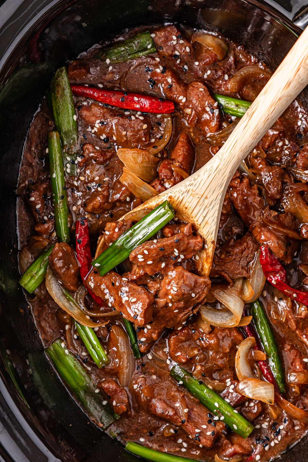 **Σιγομαγειρεμένο Μογγολικό Βοδινό** 3 A wooden spoon stirs a slow cooker Mongolian beef dish with onions, green onions, red chilies, and sesame seeds in a rich brown sauce.