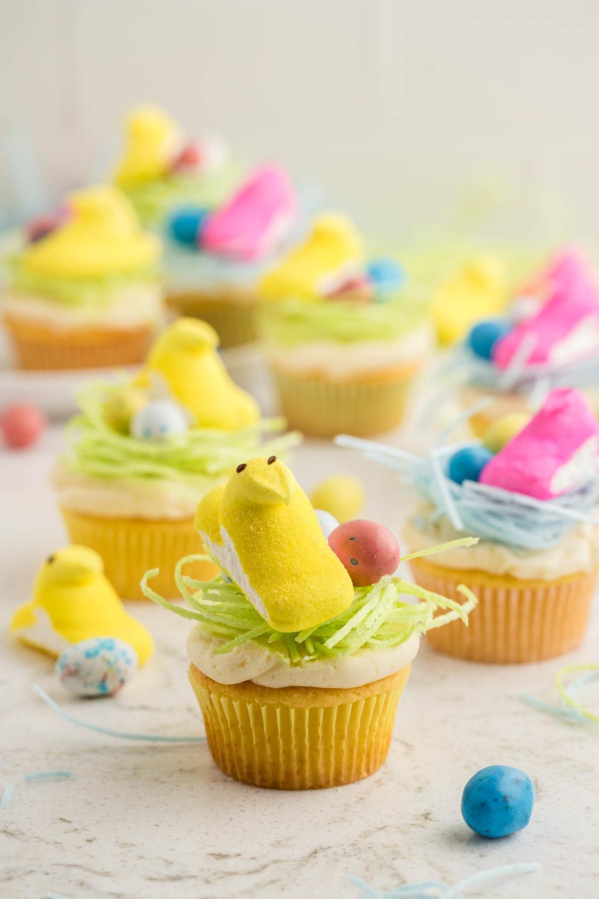 Πασχαλινά Κάπκεϊκς Πουλάκια 3 Cupcakes στολισμένα με κίτρινα μαρσμέλοου κοτόπουλα, φωλιές από πράσινη τριμμένη καρύδα και πολύχρωμα ζαχαρωτά αυγά. Άλλα διακοσμημένα cupcakes και ζαχαρωτά είναι θολά στο παρασκήνιο.