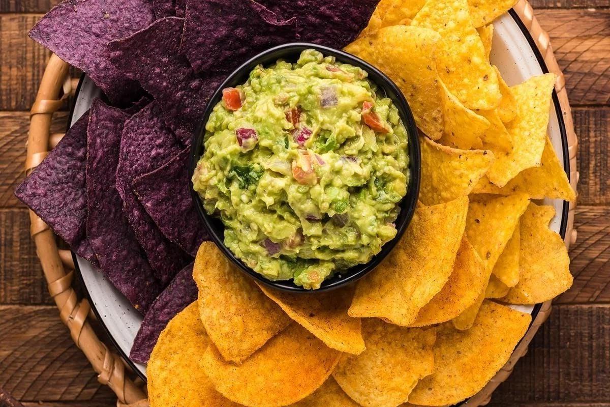 Ένα μπολ με guacamole περιτριγυρισμένο από κίτρινα και μωβ τσιπς τορτίγιας, ιδανικό για dips για πάρτι.