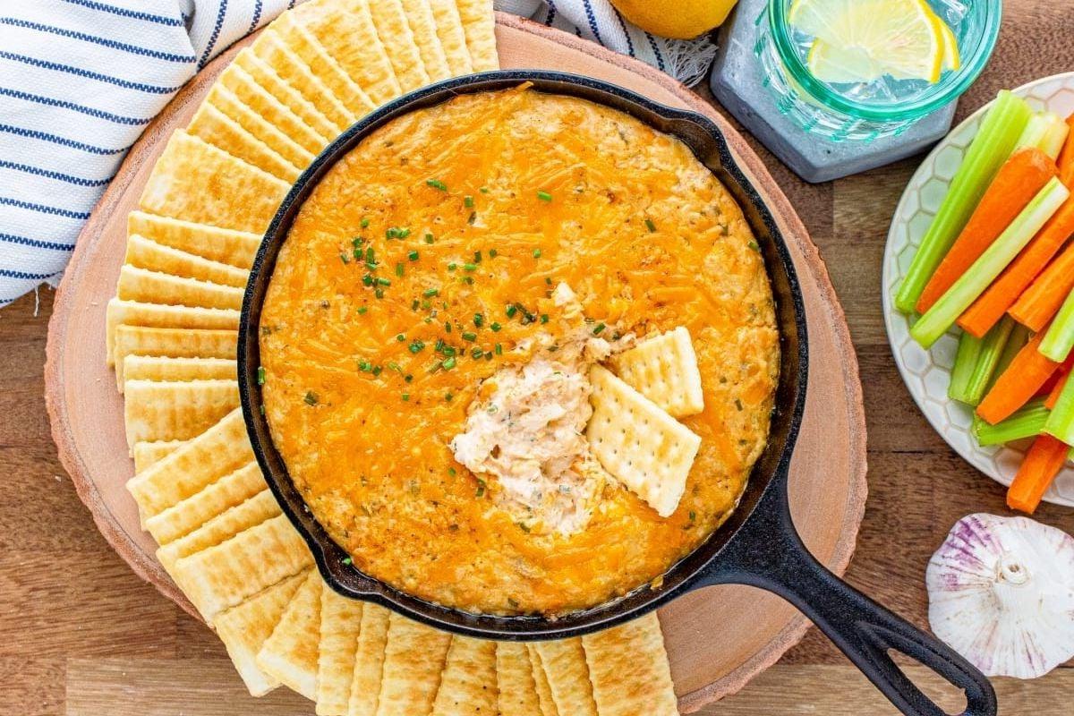 Ένα τηγάνι με ζεστό, αφράτο τυρένιο dip για πάρτι, σερβιρισμένο με διάφορα κράκερς και φρέσκα λαχανικά στο πλάι.