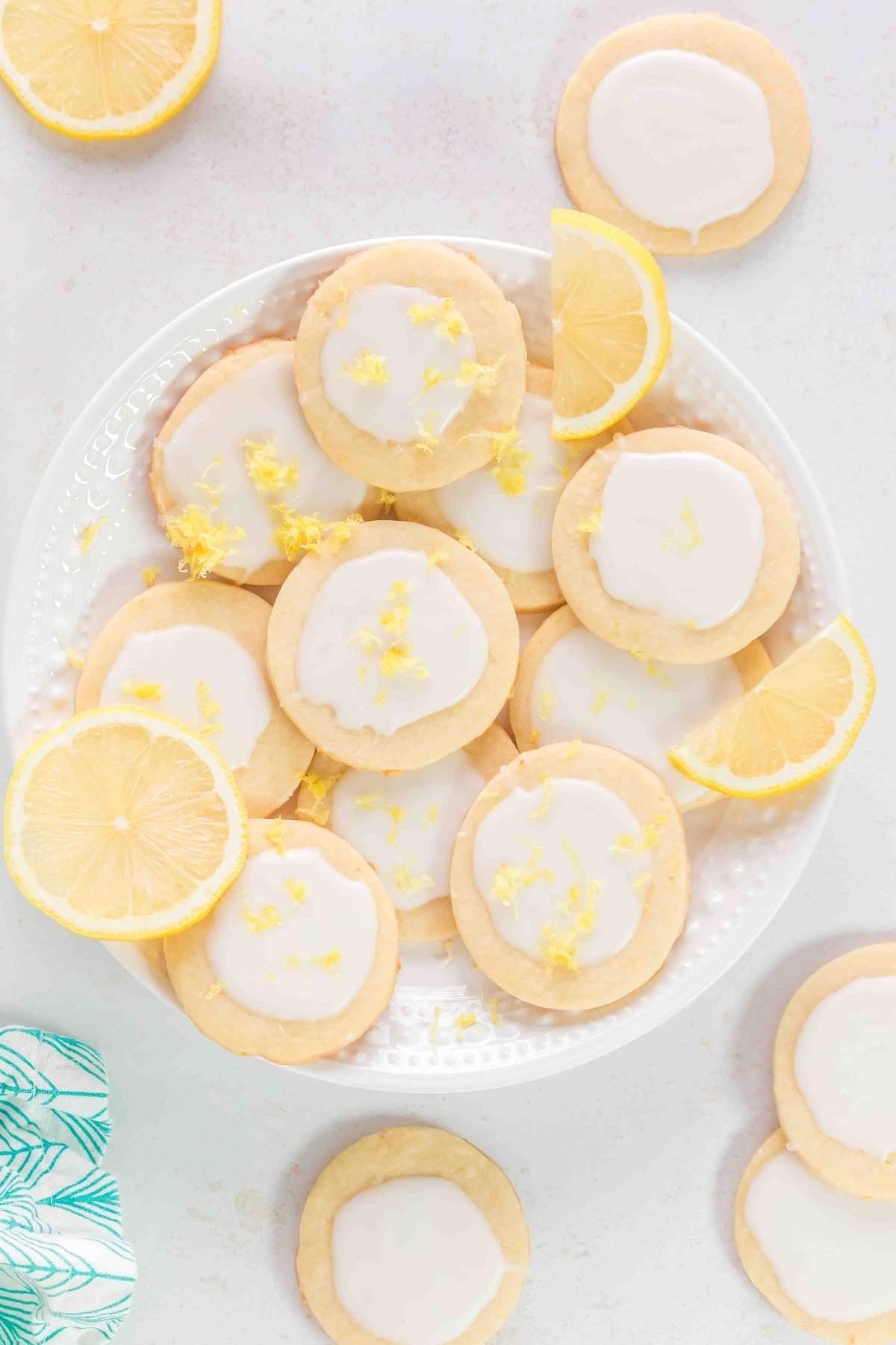 Ένα πιάτο με λεμονάτα μπισκότα shortbread με λευκό γλάσο, γαρνιρισμένα με ξύσμα λεμονιού, περιτριγυρισμένα από φέτες λεμονιού και μια γαλάζια πετσέτα με σχέδια σε λευκή επιφάνεια.