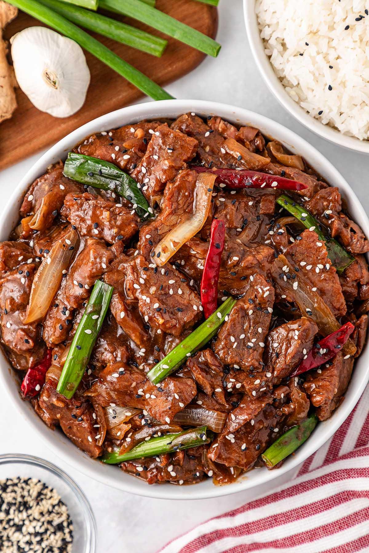**Σιγομαγειρεμένο Μογγολικό Βοδινό** 4 A bowl of stir-fried beef with green onions and red chilies, garnished with sesame seeds, reminiscent of slow cooker Mongolian beef. Rice, sliced green onions, garlic, and ginger sit invitingly in the background.
