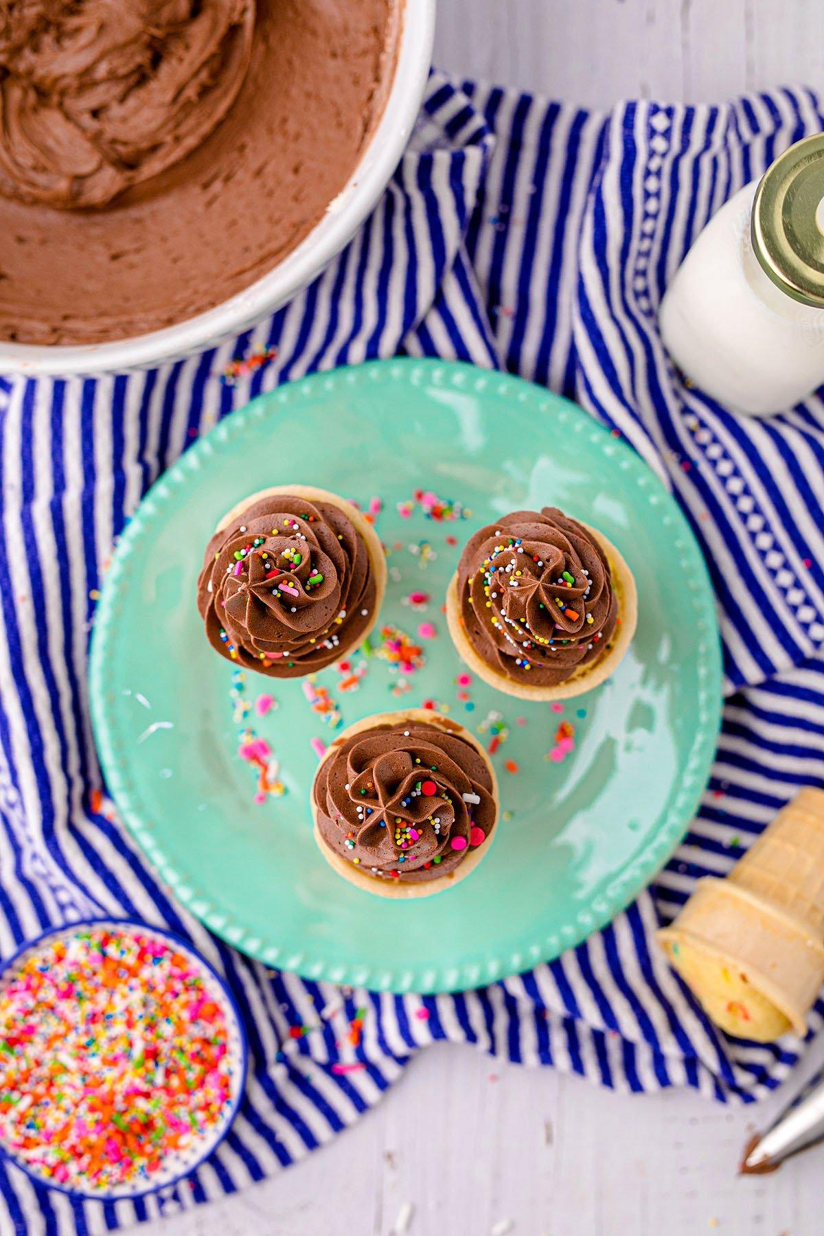 Κεκάκια Χωνάκι Παγωτού 4 Τρία cupcakes με σοκολατένια βουτυρόκρεμα και τρούφα σε ένα πράσινο πιάτο, περιτριγυρισμένα από συστατικά ψησίματος – τέλεια cupcakes σε χωνάκι παγωτού για πάρτι γενεθλίων.