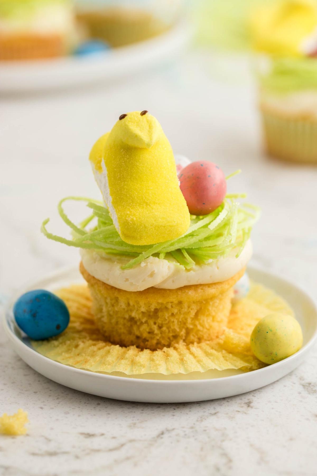 Πασχαλινά Κάπκεϊκς Πουλάκια 4 Ένα cupcake στολισμένο με ένα κίτρινο κοτόπουλο μαρσμέλοου, χρωματιστά αυγά και πράσινες διακοσμήσεις, τοποθετημένο σε ένα λευκό πιάτο.
