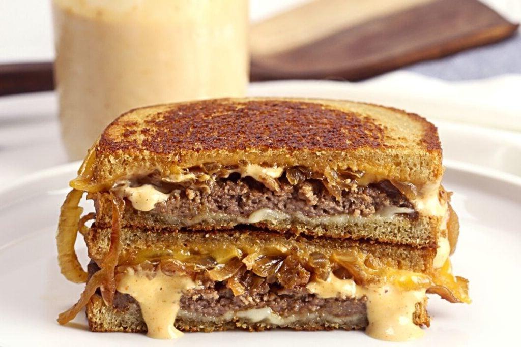 Ένα πιάτο με ένα patty melt σε ψωμί σίκαλης με καραμελωμένα κρεμμύδια, κομμένο στη μέση.