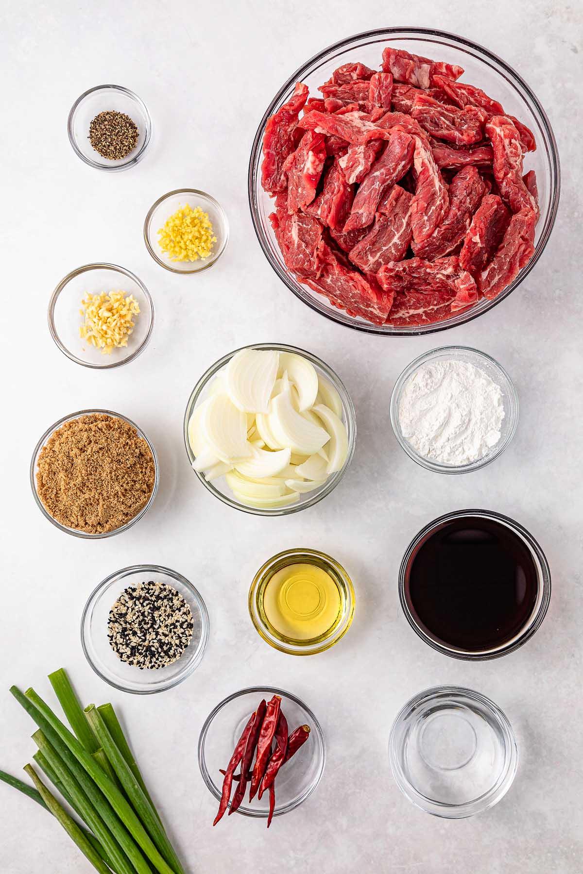 **Σιγομαγειρεμένο Μογγολικό Βοδινό** 5 Ingredients for slow cooker Mongolian beef are thoughtfully arranged: sliced beef, onions, minced garlic and ginger, ground black pepper, soy sauce, brown sugar, sesame seeds, chili peppers, cooking oil, flour, and green onions.