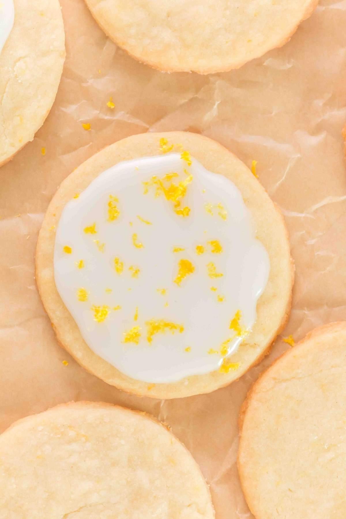 Ένα στρογγυλό λεμονάτο μπισκότο shortbread με λευκό γλάσο και μια υποψία κίτρινου ξύσματος κάθεται σε λαδόκολλα.