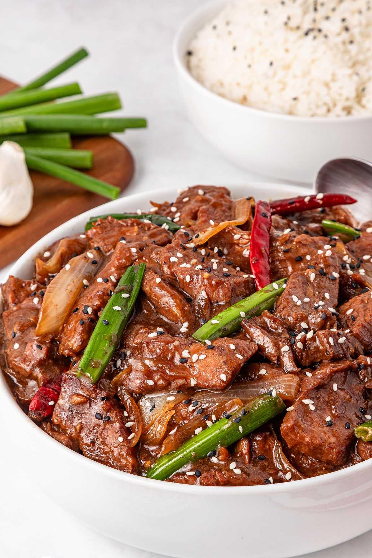 **Σιγομαγειρεμένο Μογγολικό Βοδινό** 6 A bowl of Mongolian beef stir-fry garnished with green onions, sesame seeds, and red chilies. In the background, a bowl of white rice sits alongside fresh green onions, rounding out this Asian-inspired feast.