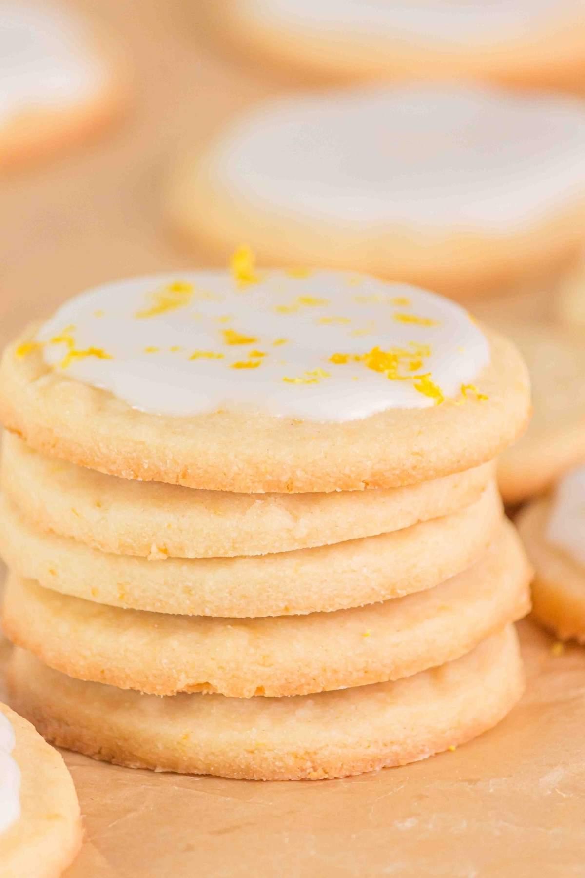Μια στοίβα από πέντε λεμονάτα μπισκότα shortbread με λευκό γλάσο και ένα πασπάλισμα ξύσματος λεμονιού στην κορυφή, τοποθετημένα σε μια επιφάνεια στρωμένη με λαδόκολλα.