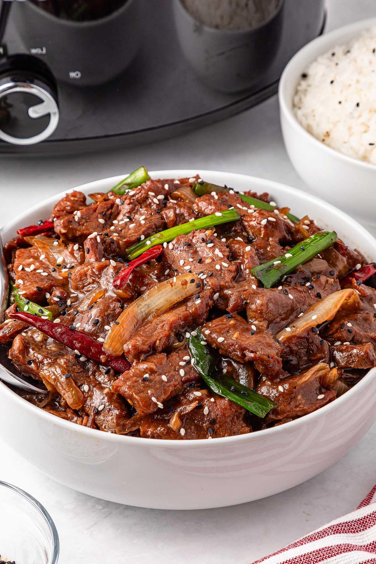 **Σιγομαγειρεμένο Μογγολικό Βοδινό** 7 A bowl of slow cooker Mongolian beef with green onions and red chilies, sprinkled with sesame seeds. A bowl of rice sits in the background.