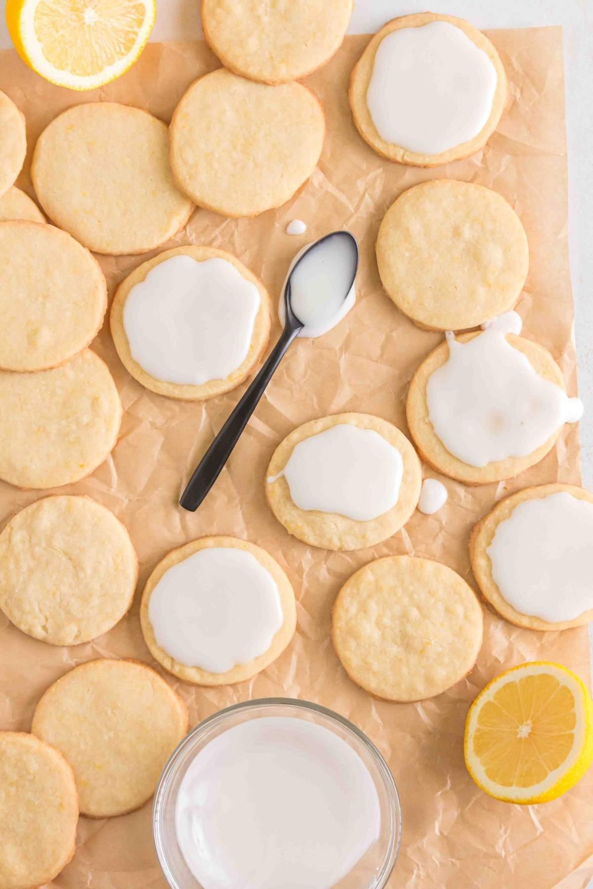 Λεμονάτα μπισκότα shortbread με λευκό γλάσο τοποθετημένα σε αντικολλητικό χαρτί, συνοδευόμενα από φρέσκες φέτες λεμονιού και ένα κουτάλι.