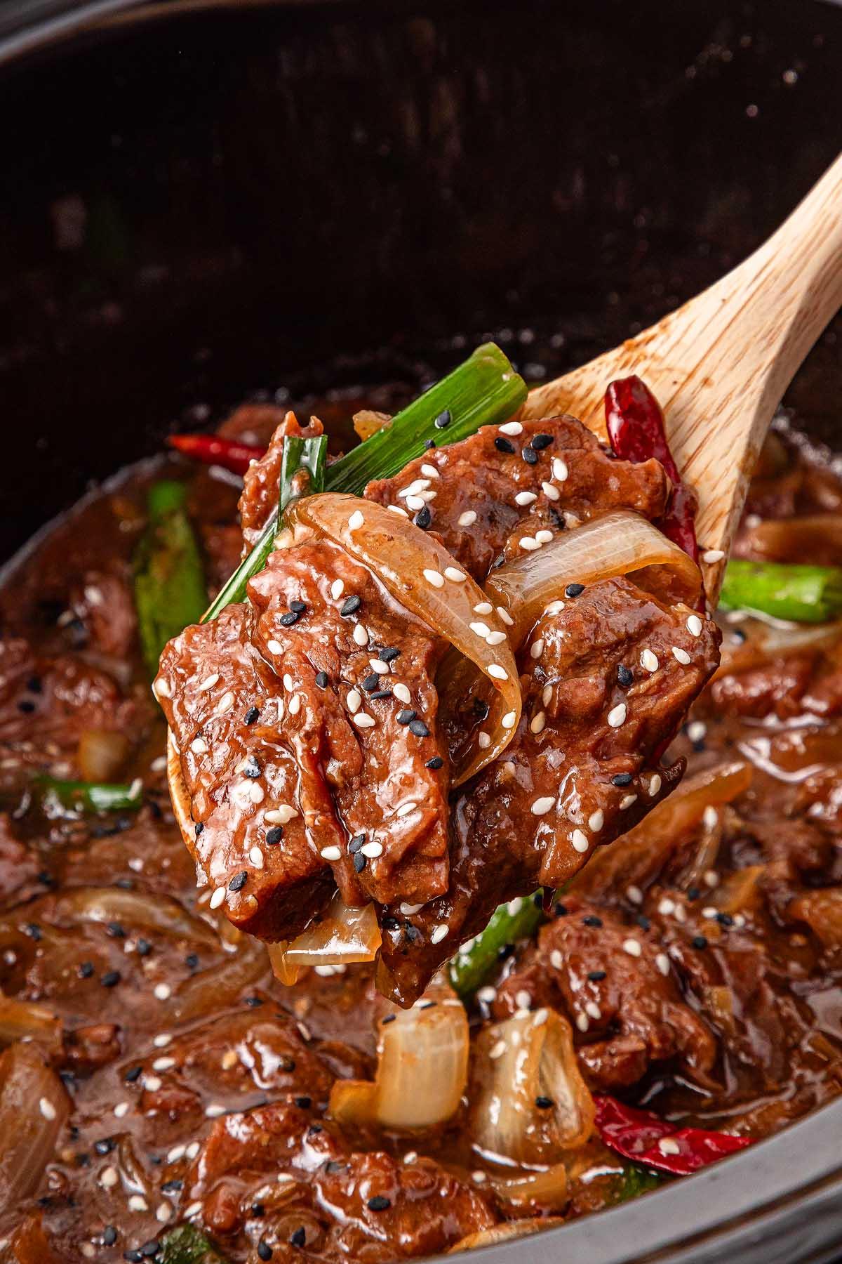 **Σιγομαγειρεμένο Μογγολικό Βοδινό** 8 A wooden spoon cradles a portion of slow cooker Mongolian beef stew with onions, green peppers, and sesame seeds. The dish looks rich and saucy, while a crockpot brimming with more savory stew waits in the background.