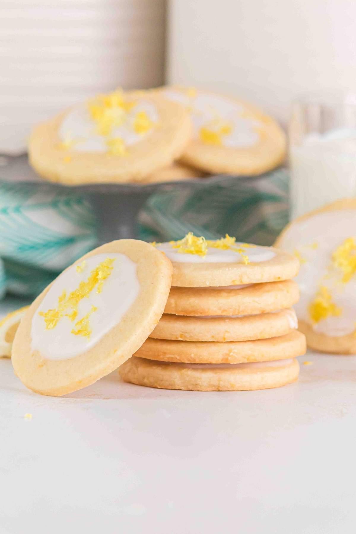 Στοίβα από λεμονάτα μπισκότα shortbread με γλάσο, γαρνιρισμένα με ξύσμα λεμονιού, σε μαρμάρινη επιφάνεια. Περισσότερα μπισκότα και ένα ποτήρι γάλα βρίσκονται στο παρασκήνιο.