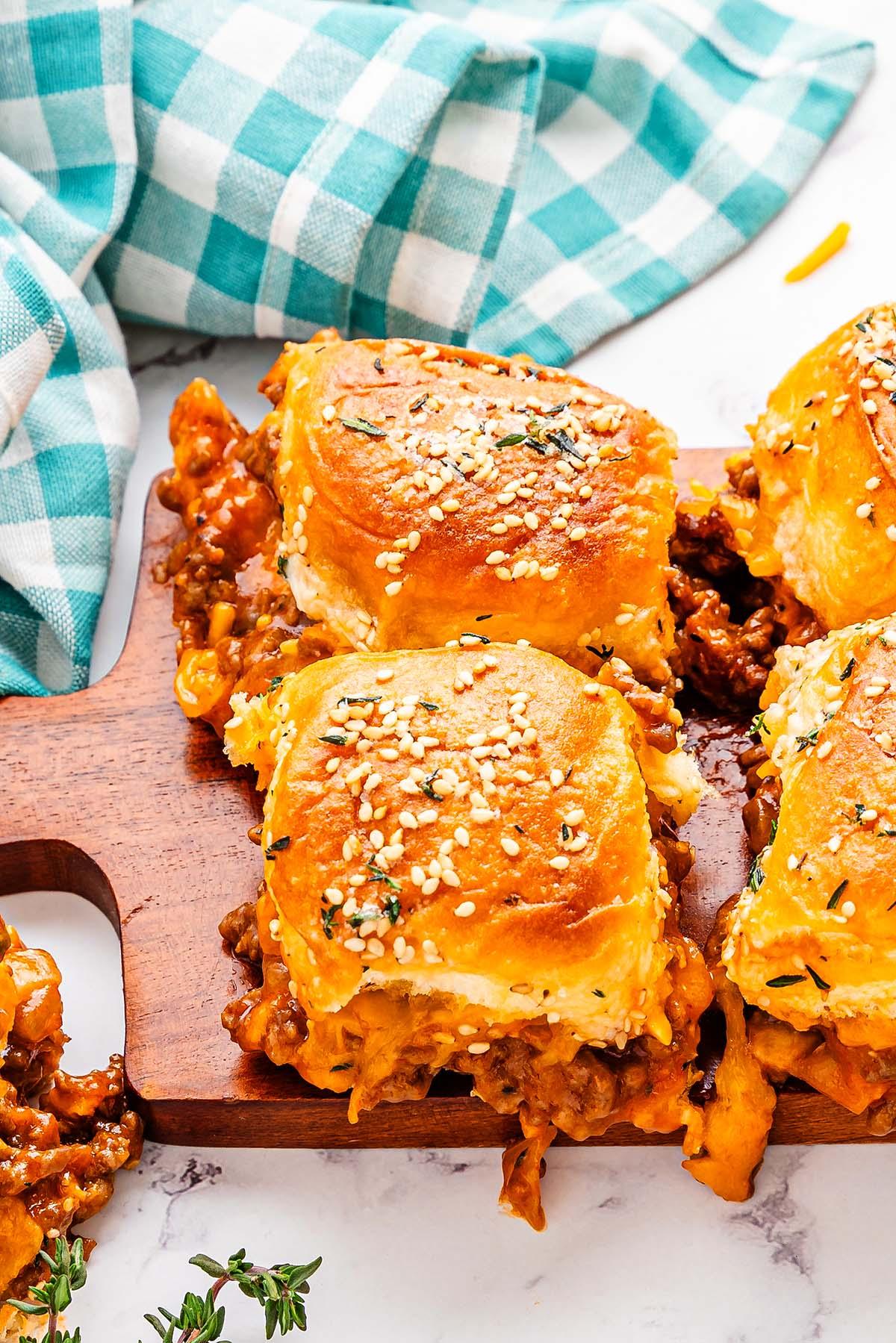 Σλόπι Τζο Μίνι Σάντουιτς 9 Sliders με ψωμάκια με σουσάμι γεμισμένα με ένα αλμυρό μείγμα Sloppy Joe και τυρί, τοποθετημένα σε ξύλινη σανίδα δίπλα σε ένα μπλε και λευκό καρό ύφασμα.