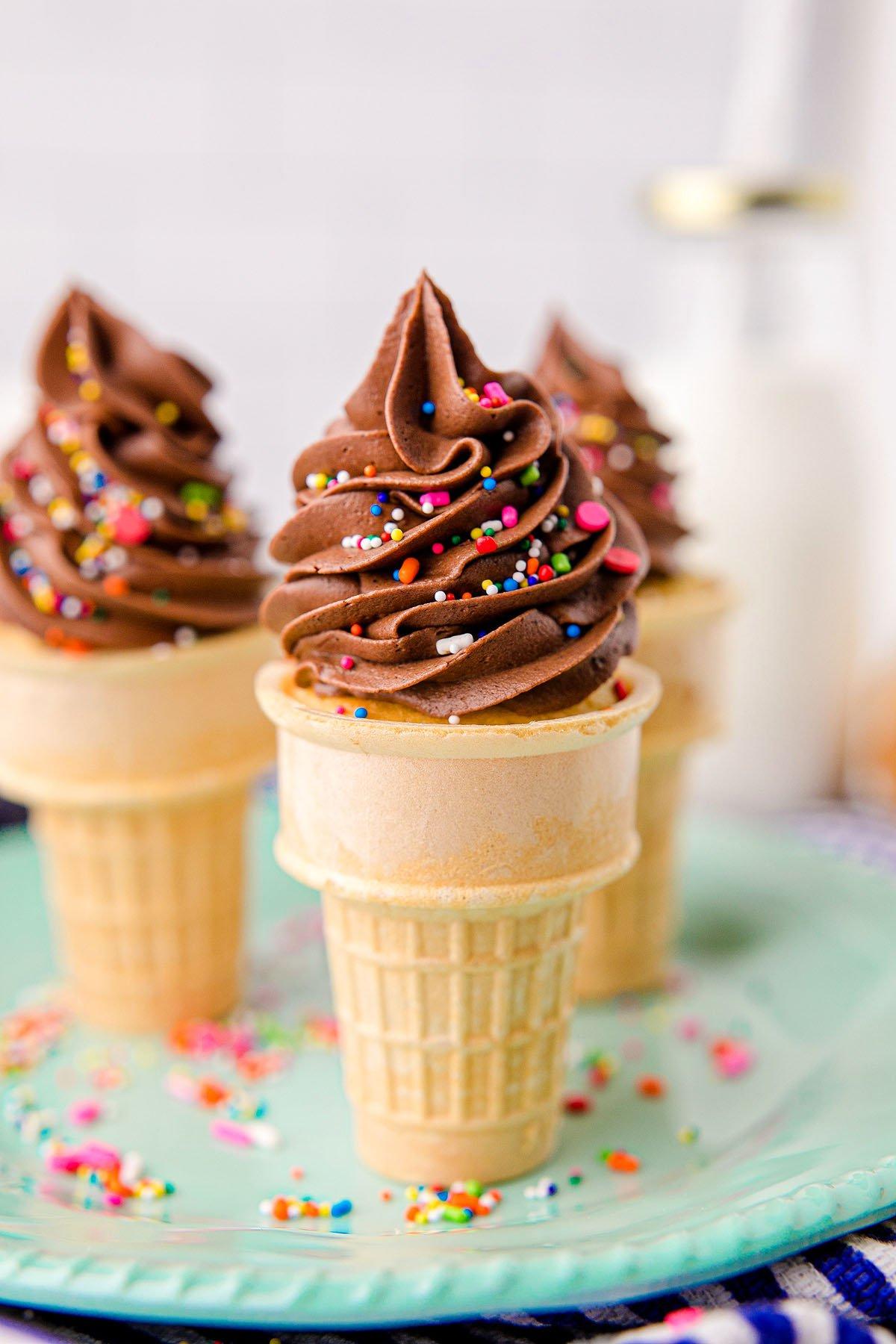 Κεκάκια Χωνάκι Παγωτού 9 Cupcakes σε χωνάκι παγωτού με σοκολατένια βουτυρόκρεμα και πολύχρωμη τρούφα αποτελούν μια διασκεδαστική λιχουδιά, ιδανικά ως cupcakes για πάρτι γενεθλίων, σερβιρισμένα σε ένα ανοιχτό πράσινο πιάτο.