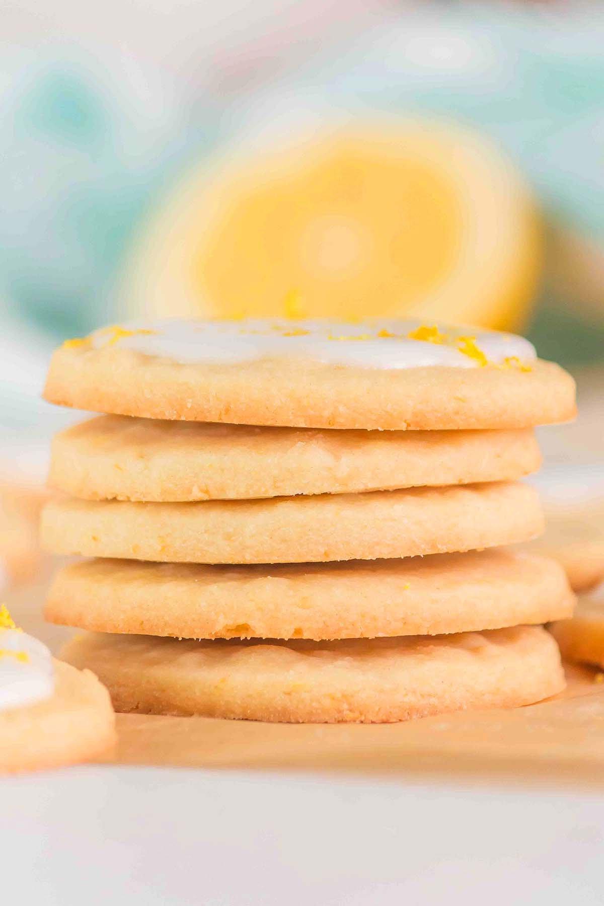 Μια στοίβα από λεμονάτα μπισκότα shortbread με γλάσο ξεκουράζεται φιλόξενα σε λαδόκολλα. Ένα κομμένο λεμόνι είναι θολό στο παρασκήνιο, ενισχύοντας την πιπεράτη ελκυστικότητα.