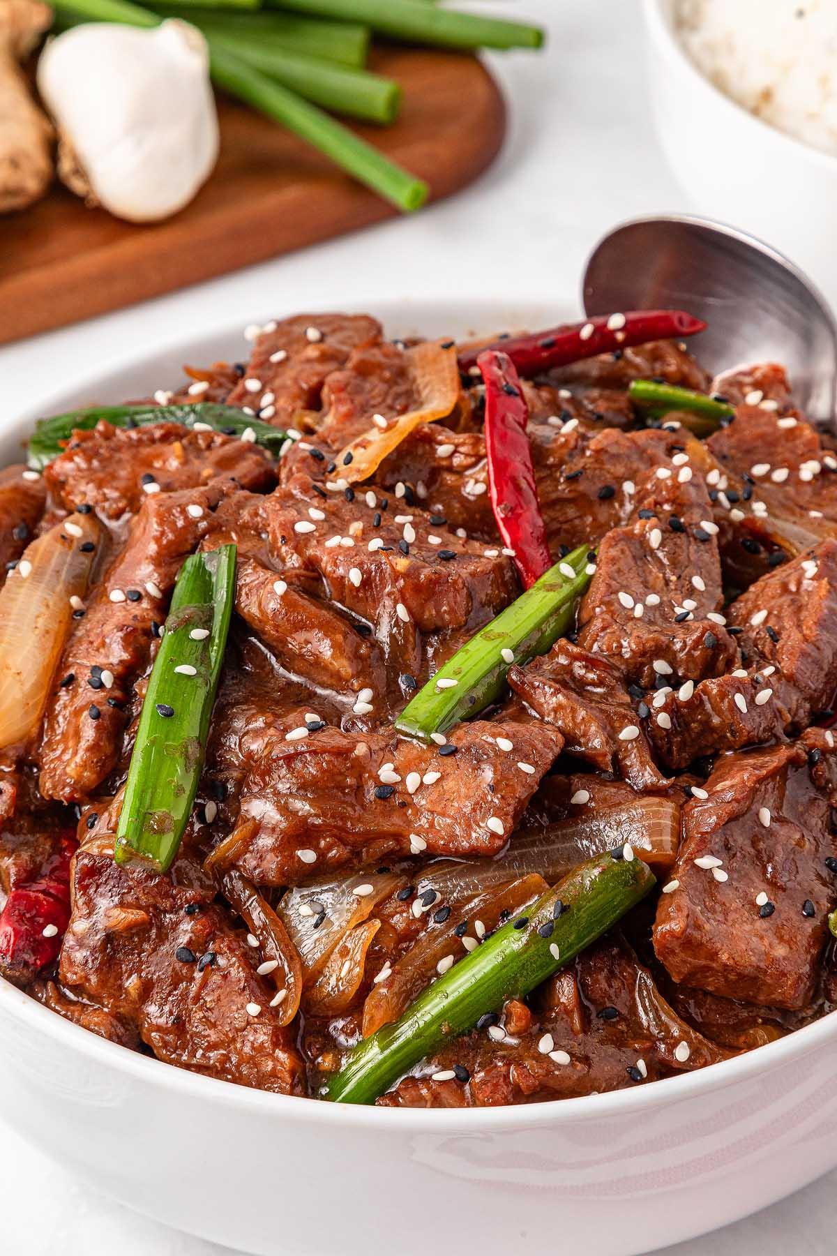 **Σιγομαγειρεμένο Μογγολικό Βοδινό** 10 A bowl of spicy slow-cooker Mongolian beef stir-fry with green onions, red chili peppers, and sesame seeds. A spoon rests inside the bowl. Garlic and more green onions are on a wooden board in the background.