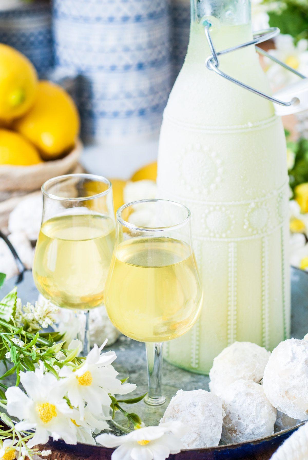 Συνταγή Ιταλικού Λιμοντσέλο 10 Δύο ποτήρια limoncello κοσμούν έναν δίσκο δίπλα σε ένα διακοσμητικό μπουκάλι και λευκά λουλούδια, αναδεικνύοντας μια ιταλική συνταγή limoncello. Στο φόντο, λεμόνια και μπουκάλια με σχέδια προσθέτουν μια νότα κομψότητας.