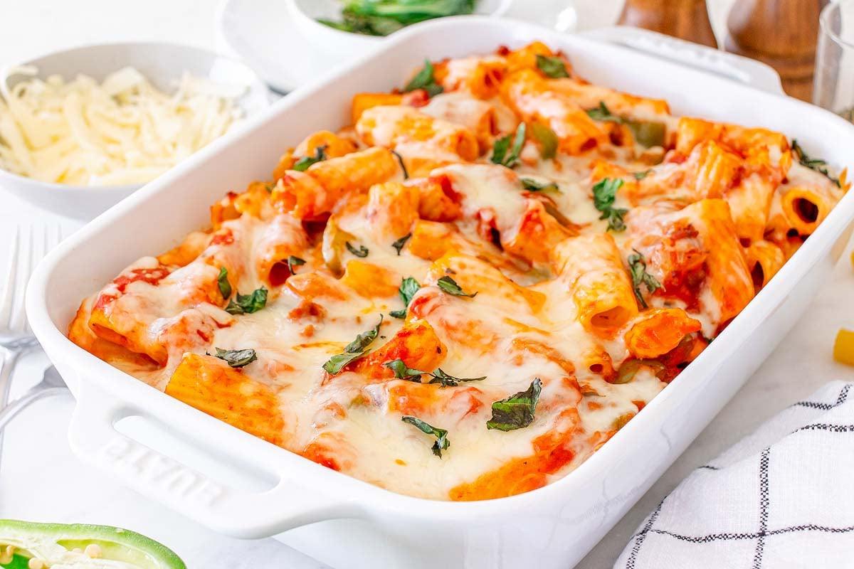 Πάστα στο φούρνο 2 Ένα πιάτο Pasta al Forno με λιωμένο τυρί και βότανα σε ένα λευκό πυρίμαχο σκεύος, με τριμμένο τυρί στο παρασκήνιο.
