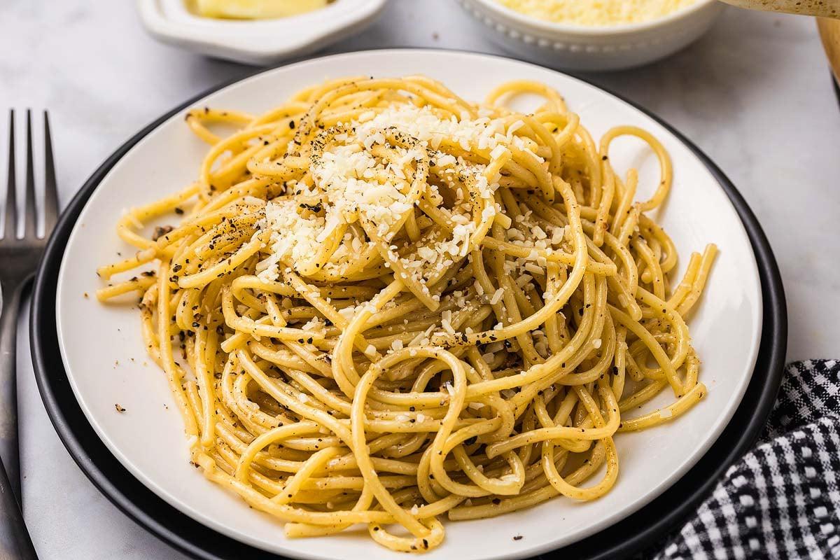 Τυρί και Πιπέρι 2 Ένα αχνιστό πιάτο cacio e pepe, στριμμένο με τριμμένο τυρί και μαύρο πιπέρι, βρίσκεται δελεαστικά στο τραπέζι, συνοδευόμενο από μια καρό πετσέτα και ένα γυαλιστερό πιρούνι.