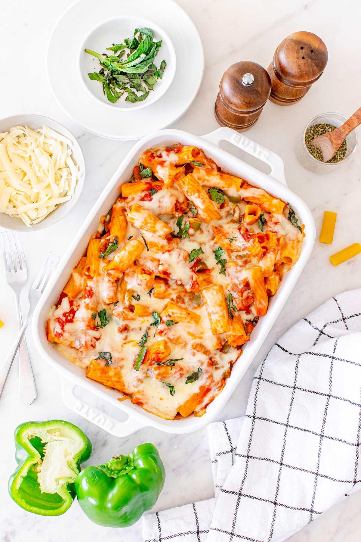 Πάστα στο φούρνο 11 Ένα ταψί τυρένιο Pasta al Forno σε ένα τραπέζι με μισές πιπεριές, τριμμένο τυρί, μια ασπρόμαυρη καρό πετσέτα, αλατοπιπεριέρες και ένα μικρό μπολ με βότανα.