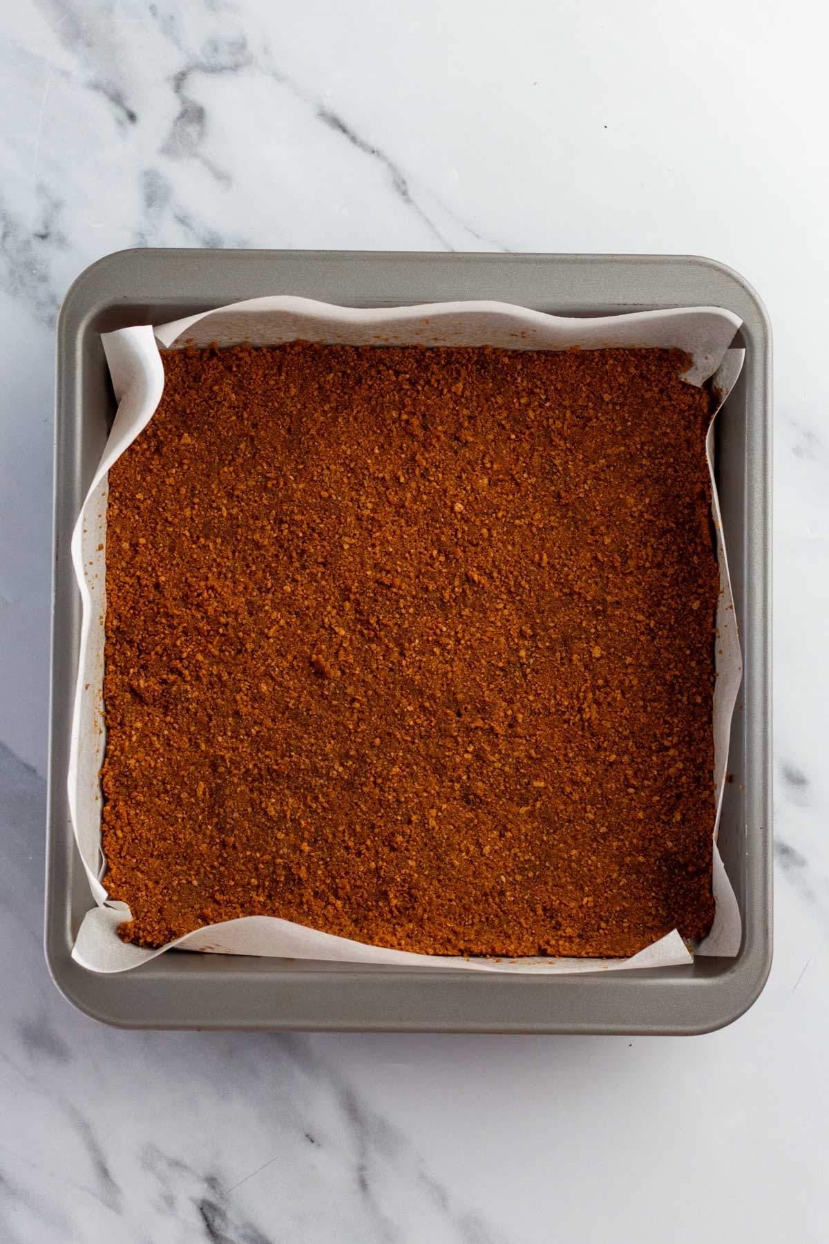 Ταψί γεμάτο με μια λεπτής υφής καφέ μείγμα s'mores brownie, καλυμμένο με αντικολλητικό χαρτί, τοποθετημένο σε λευκή μαρμάρινη επιφάνεια.