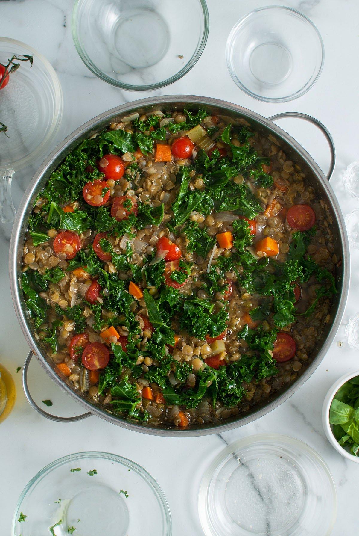 Ιταλική Σούπα Φακής 17 Μια κατσαρόλα Zuppa Di Lenticchie, μια ιταλική σούπα φακής που ξεχειλίζει από φρέσκα λαχανικά όπως λάχανο κέιλ, ντοματίνια, φακές και καρότα, βρίσκεται περιτριγυρισμένη από άδεια γυάλινα μπολ.