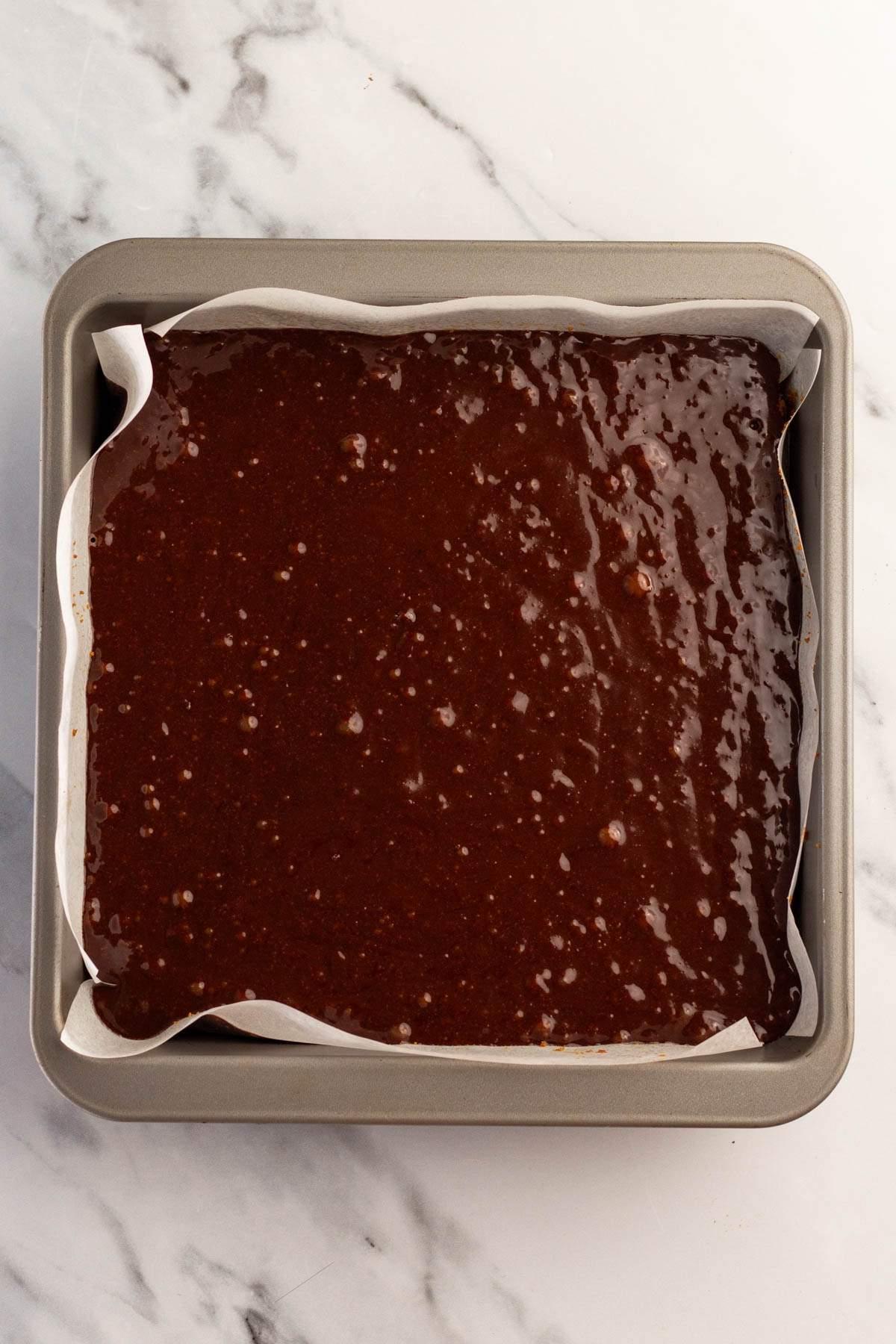 Ένα τετράγωνο ταψί γεμάτο με ωμή ζύμη s'mores brownie, στρωμένο με αντικολλητικό χαρτί σε ένα μαρμάρινο πάγκο κουζίνας.