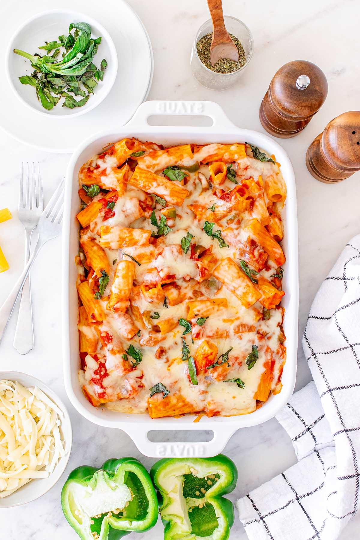 Πάστα στο φούρνο 4 Μια ιταλική συνταγή Pasta al Forno με λιωμένο τυρί και βασιλικό σε ένα λευκό πυρίμαχο σκεύος, περιτριγυρισμένο από πράσινες πιπεριές, τριμμένο τυρί, μαχαιροπίρουνα και μπαχαρικά σε έναν μαρμάρινο πάγκο.