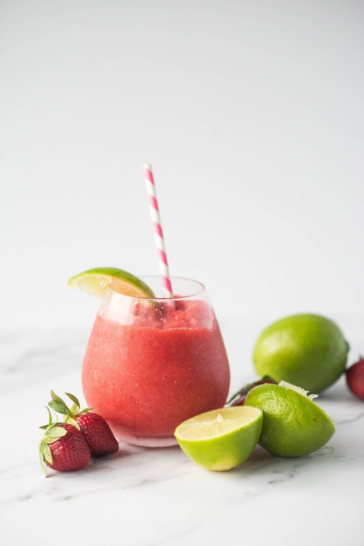 Ένα ποτήρι κόκκινο smoothie με ριγέ καλαμάκι, φέτα λάιμ, λάιμ και φράουλες σε λευκή επιφάνεια — ένας εύκολος τρόπος να φτιάξετε ένα ντάικιρι ή να δοκιμάσετε την καλύτερη συνταγή ντάικιρι για μια δροσιστική ανατροπή.