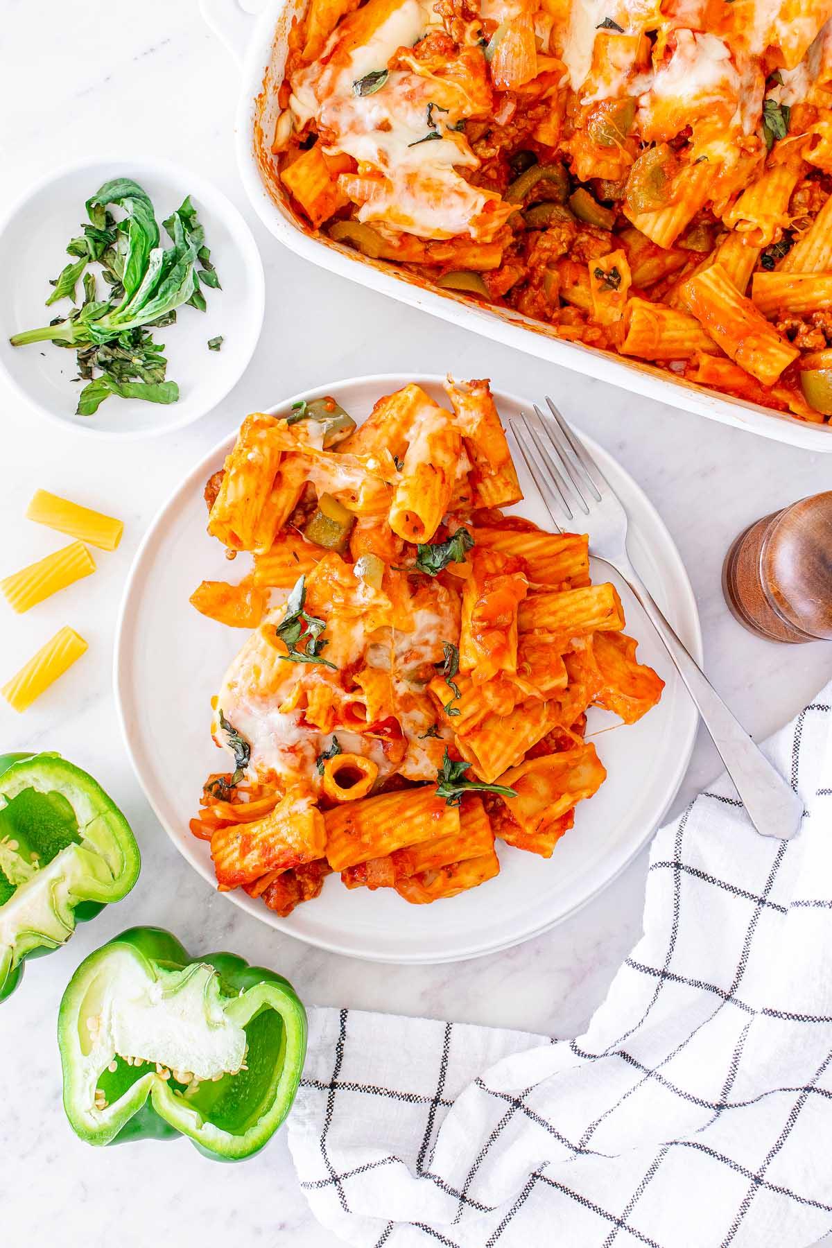 Πάστα στο φούρνο 7 Ένα πιάτο Pasta al Forno με ψητά ριγκατόνι και λιωμένο τυρί, σερβιρισμένο με φρέσκο βασιλικό και πράσινες πιπεριές, δίπλα σε ένα λευκό μπολ που περιέχει επιπλέον βασιλικό και μισή πράσινη πιπεριά.