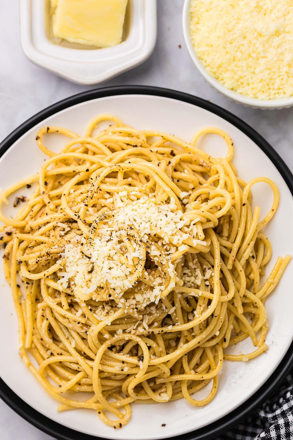 Τυρί και Πιπέρι 7 Ένα πιάτο cacio e pepe περιλαμβάνει σπαγγέτι καρυκευμένα με μαύρο πιπέρι και τριμμένο τυρί. Δίπλα στο πιάτο, ένα μπολ με τριμμένο τυρί και ένα κομμάτι βούτυρο ολοκληρώνουν τη σκηνή.