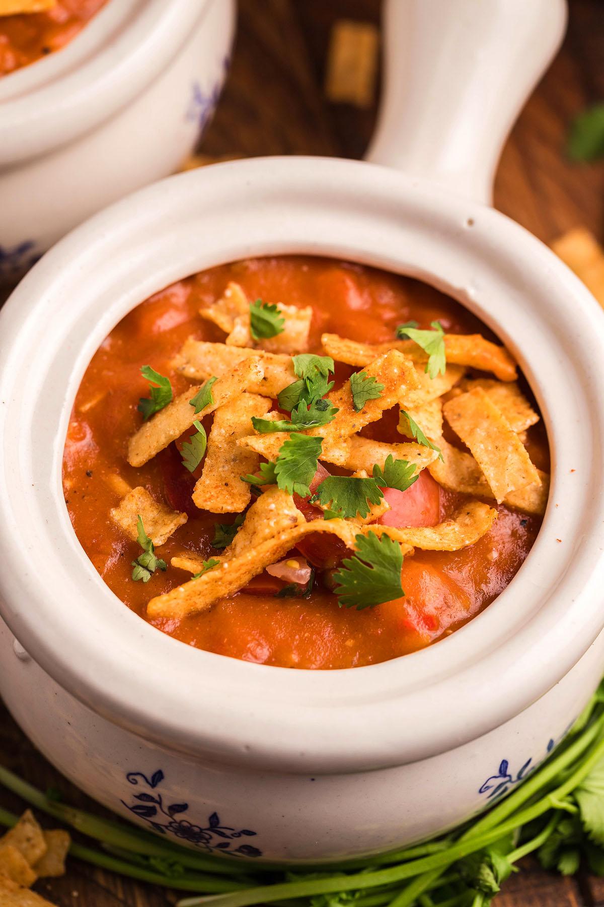 Σούπα Εντσιλάδα Κοτόπουλου – Τύπου Chili's 7 Ένα μπολ με μεξικάνικη σούπα κοτόπουλου, εμπλουτισμένο με τρυφερό κοτόπουλο σούβλας και γαρνιρισμένο με λωρίδες τορτίγιας και κόλιανδρο, βρίσκεται δελεαστικά σε μια λευκή κεραμική κατσαρόλα σε μια ξύλινη επιφάνεια.