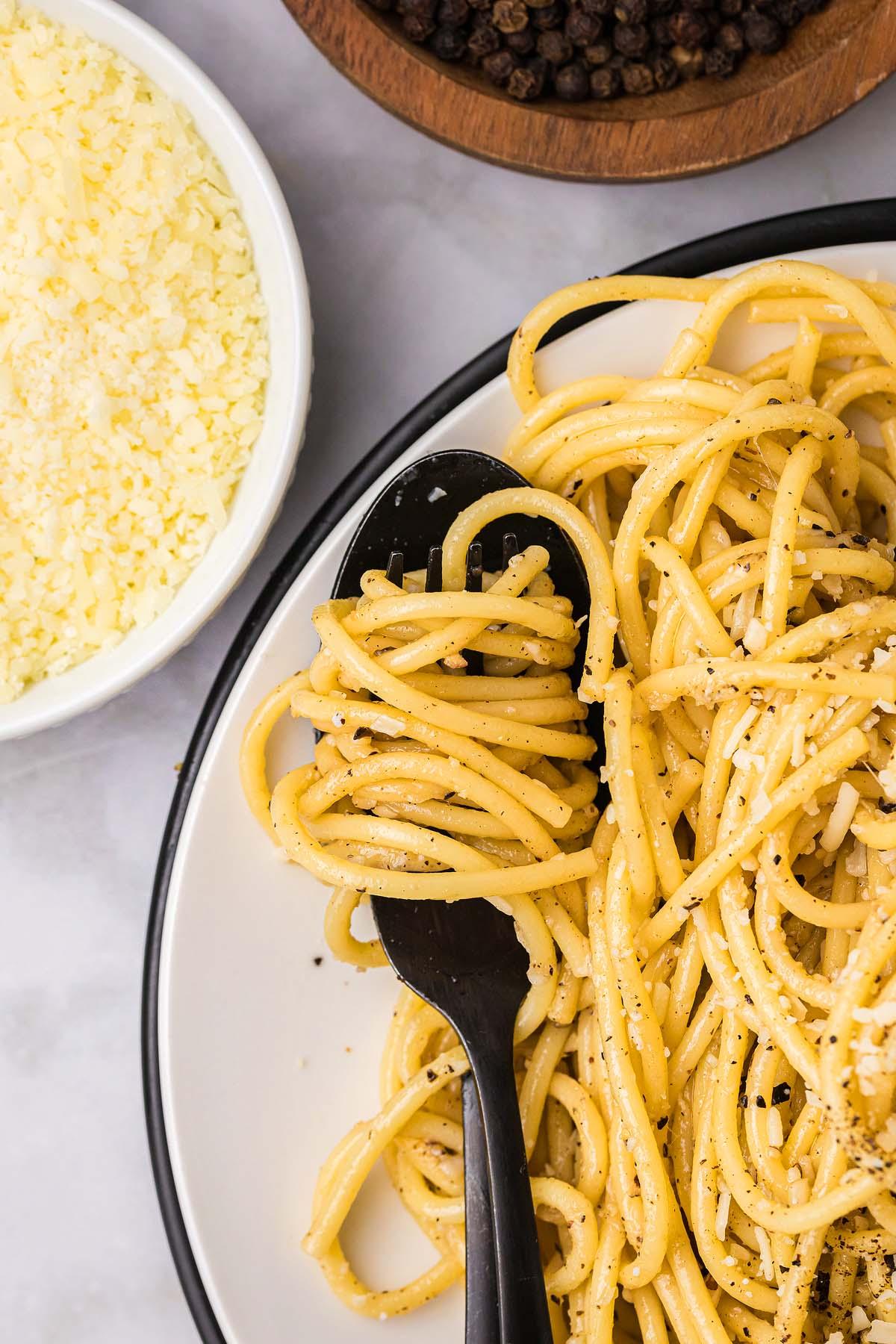 Τυρί και Πιπέρι 8 Ένα πιάτο cacio e pepe, καρυκευμένο με μαύρο πιπέρι και τυρί, περιλαμβάνει noodles κομψά τυλιγμένα γύρω από ένα μαύρο πιρούνι. Κοντά, ένα μπολ με τριμμένο τυρί και ολόκληρους κόκκους πιπεριού ενισχύουν τη μινιμαλιστική γοητεία αυτού του κλασικού πιάτου.