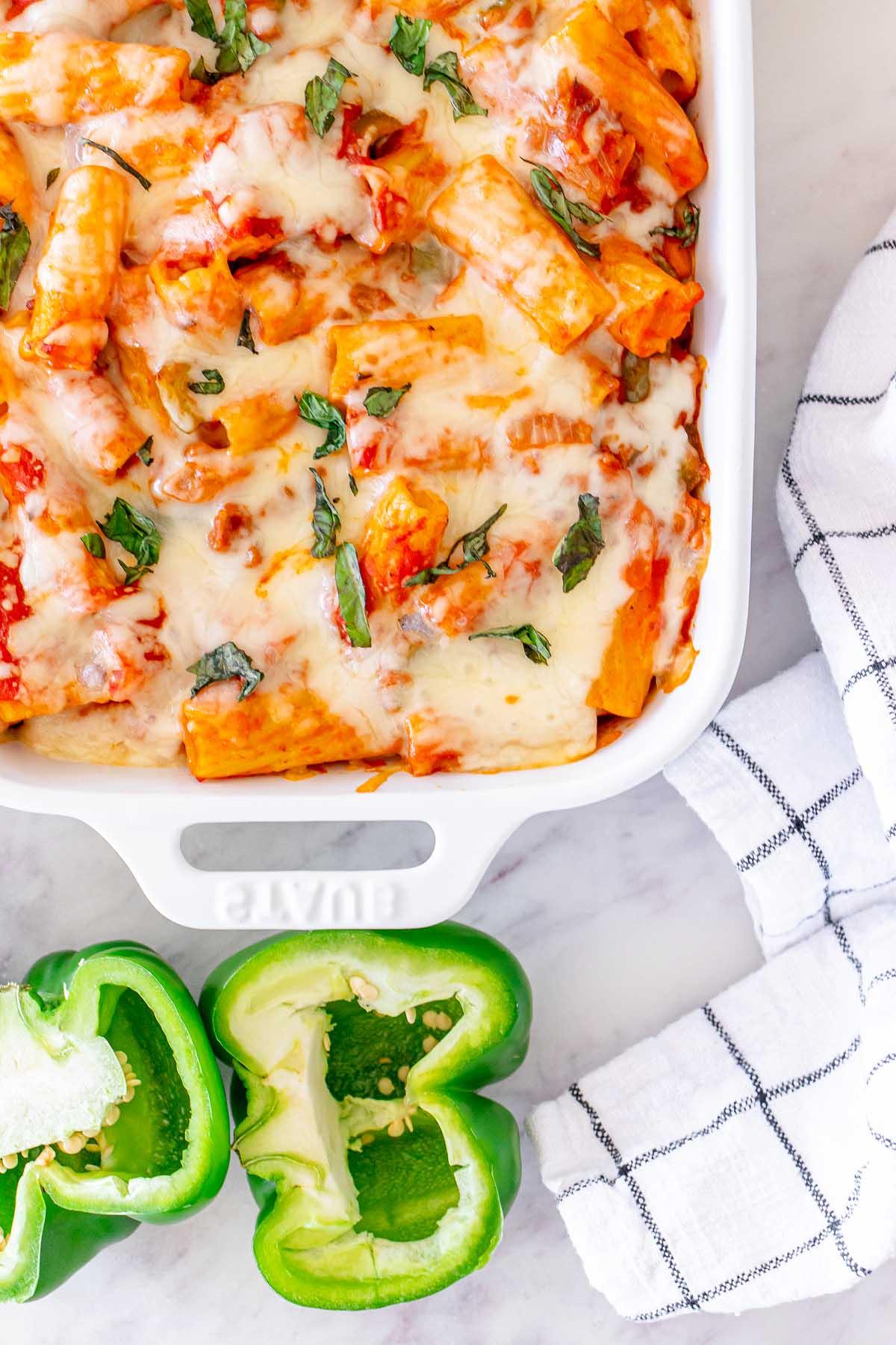 Πάστα στο φούρνο 10 Αυθεντικό pasta al forno με λιωμένο τυρί και φρέσκα βότανα σε ένα λευκό ταψί δίπλα σε μια κομμένη πράσινη πιπεριά και ένα ριγέ ύφασμα.