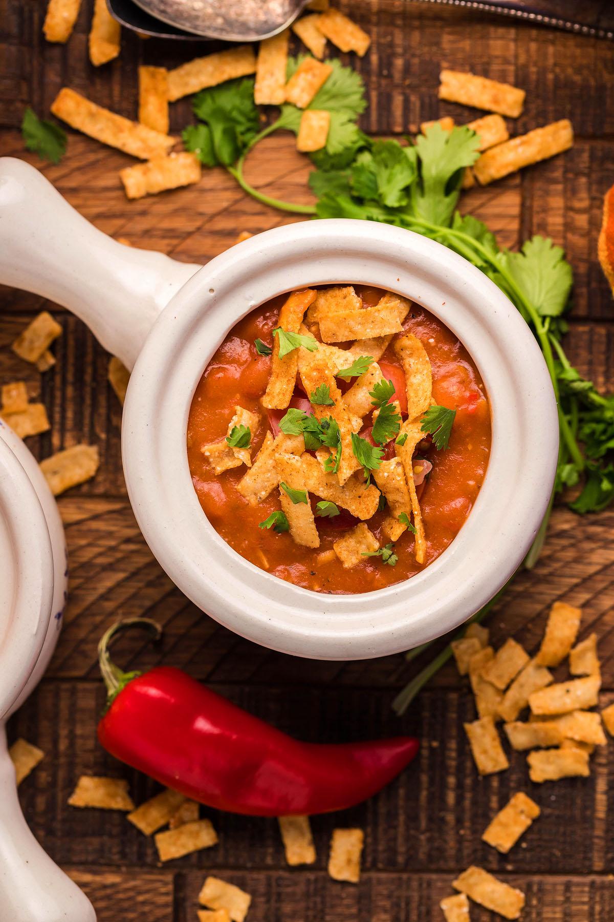 Σούπα Εντσιλάδα Κοτόπουλου – Τύπου Chili's 10 Ένα μπολ με σούπα κοτόπουλου εντσιλάδας γαρνιρισμένο με τραγανές λωρίδες και κόλιανδρο βρίσκεται σε ένα ξύλινο τραπέζι, συνοδευόμενο από μια κόκκινη πιπεριά τσίλι και σκορπισμένες λωρίδες. Αυτή η συνταγή Chili's Copycat φέρνει μια γεύση χαράς στο τραπέζι σας.