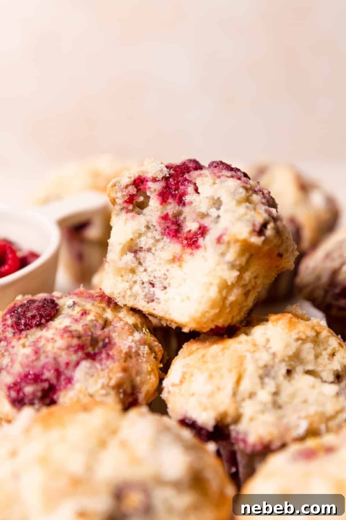 A pile of moist lemon raspberry muffins with the top one missing a bite to show the fluffy interior and juicy raspberries.